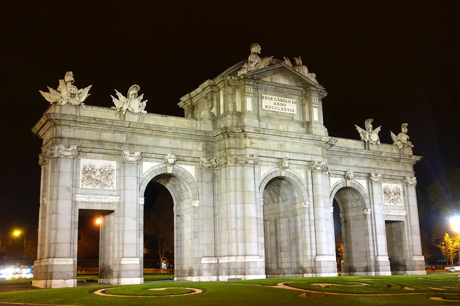 The Puerta de Alcalá lit at night, a pale stone gate with three arches and sculpted figures above, set in a garden roundabout