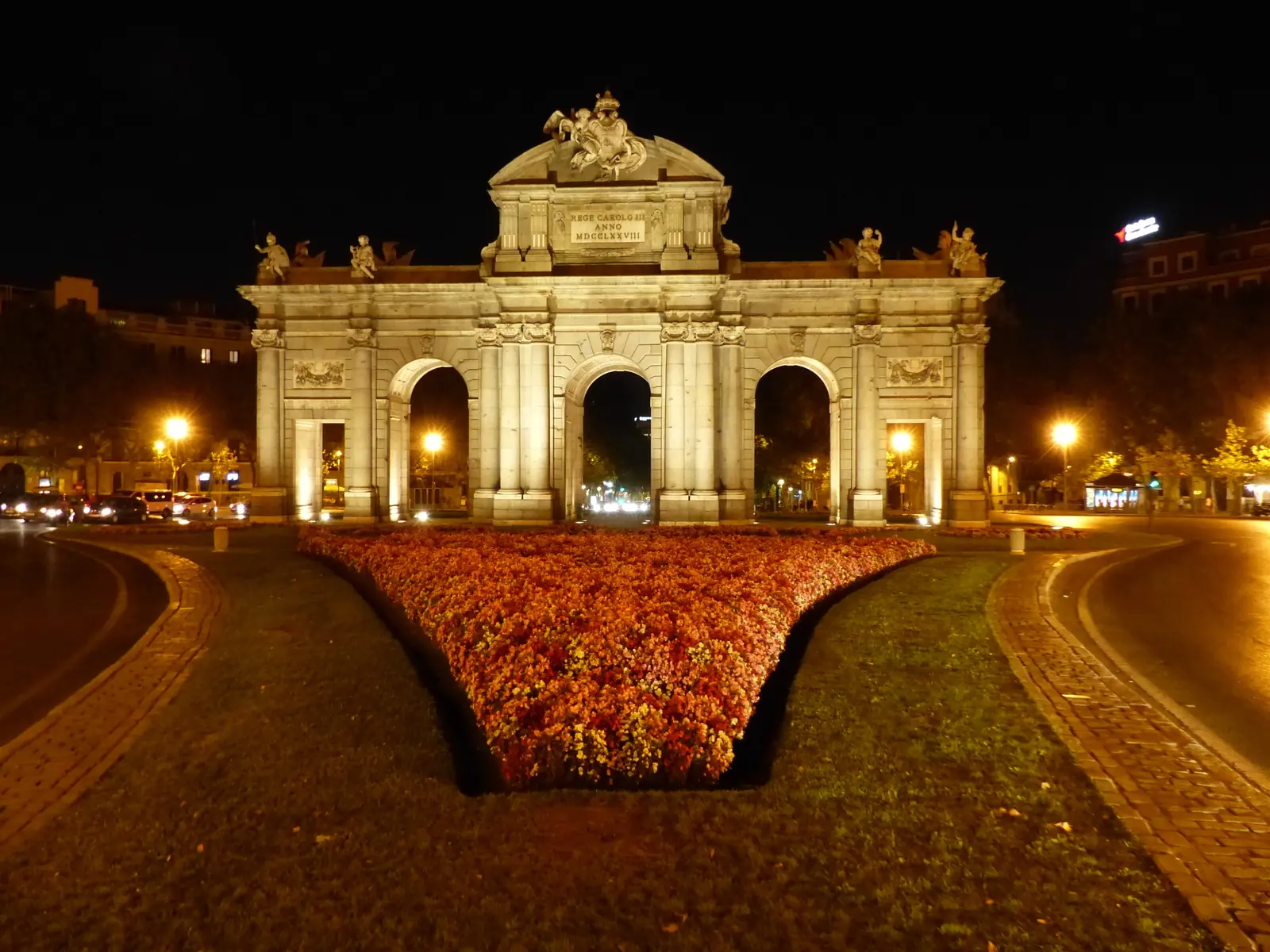 The Puerta de Alcalá lit at night from a low angle, with a bed of orange flowers in the foreground and a road curving around the roundabout