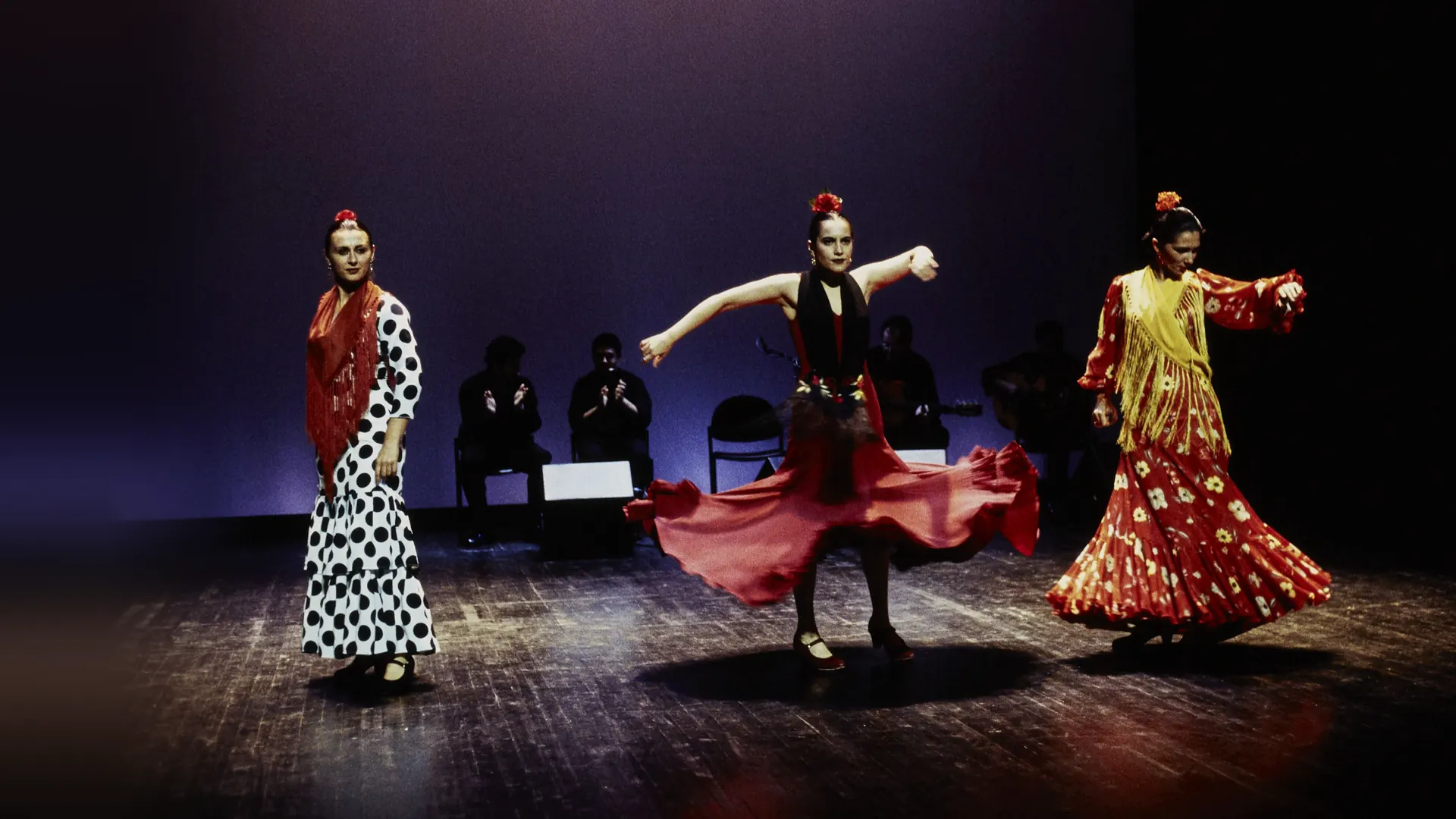 Three flamenco dancers on a dark stage under violet light: a woman in a polka-dot dress, a woman in a red skirt mid-turn and a woman in a yellow shawl, with seated musicians behind them