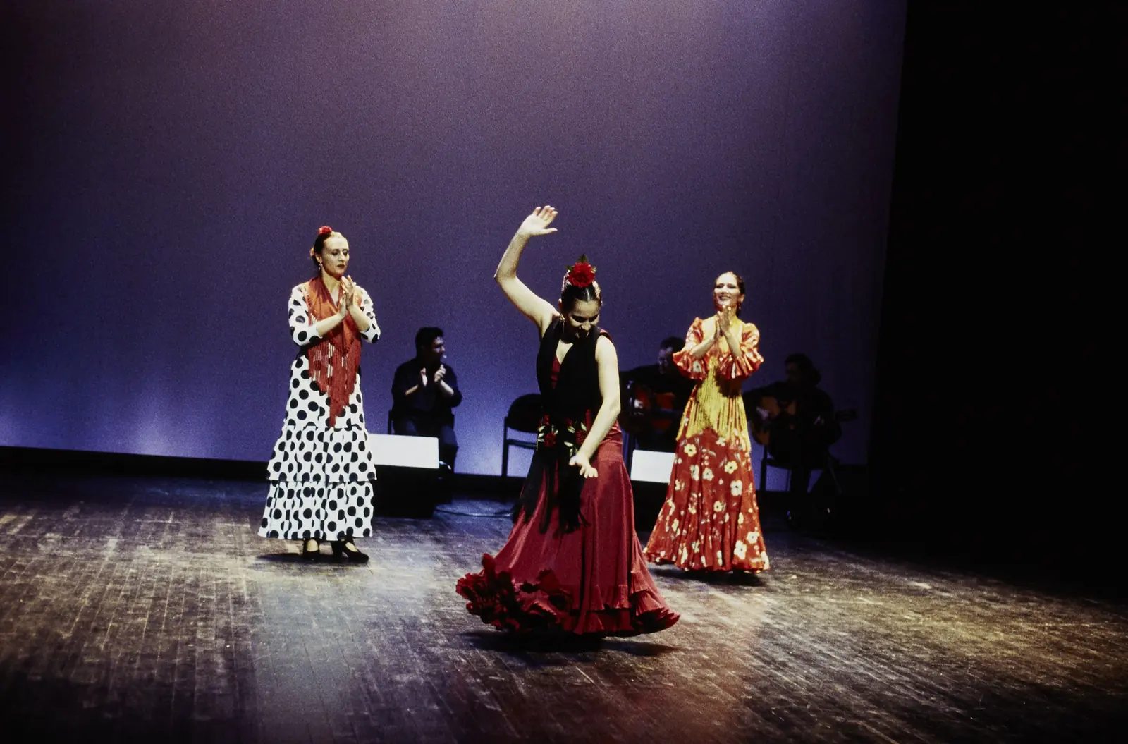 A flamenco dancer in a red skirt with one arm raised on a dark stage under violet light, with two other dancers in polka-dot and yellow dresses beside her