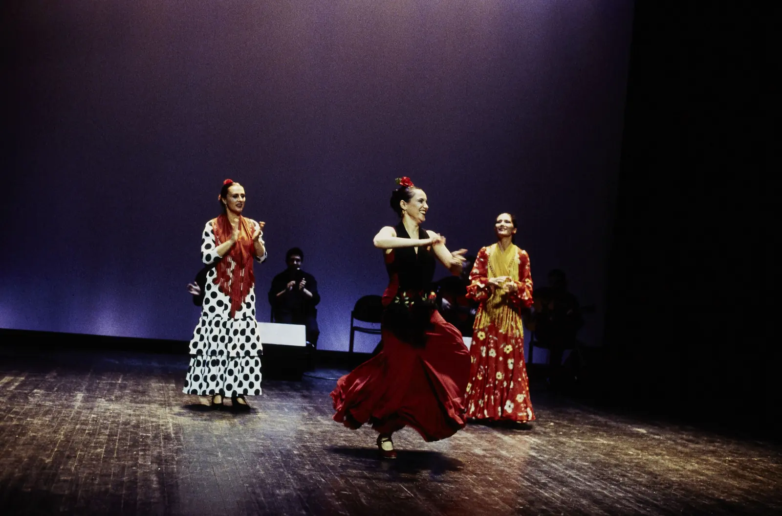Three flamenco dancers in polka-dot and red dresses on a dark stage under violet light, with seated musicians behind them