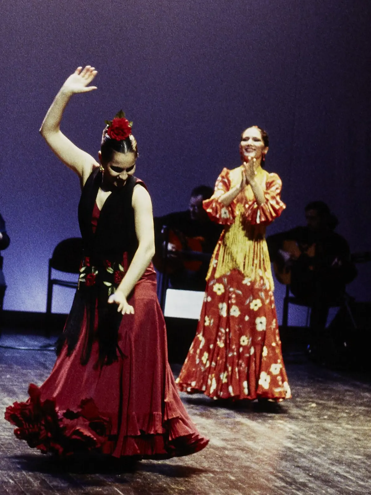 A flamenco dancer in a red skirt with one arm raised on a dark stage, and a second dancer in a yellow shawl clapping behind her